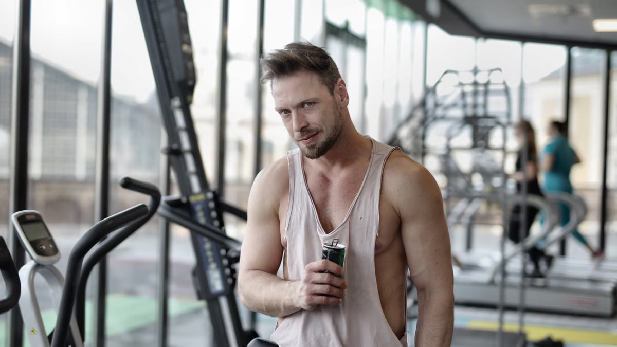 Positive young muscular bearded male athlete in casual sportswear standing near window in modern gym with bottle of cold energy drink and looking at camera